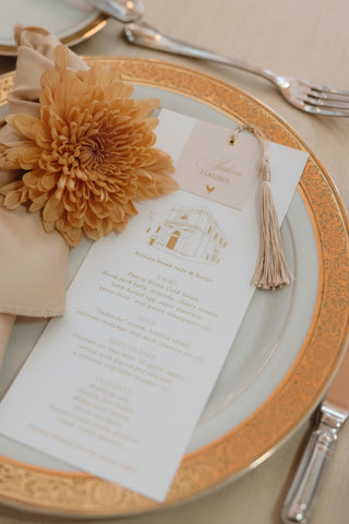Decorative table setting with a gold flower, menu, and tassel on a plate.